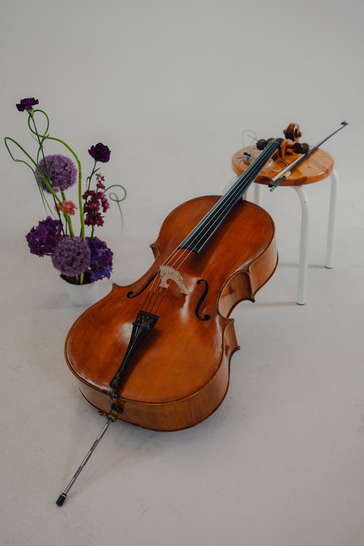 Ein Cello lehnt an einem Hocker, daneben lila Blumen
